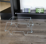 A silver metal wire display stand or shelf insert placed on a wood-look floor in front of black shelving with storage boxes on a pegboard wall.