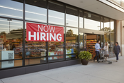 Now Hiring red vinyl banner 8 feet wide hung on the exterior window or entrance of a retail store with a welcoming hiring atmosphere and professional storefront setting