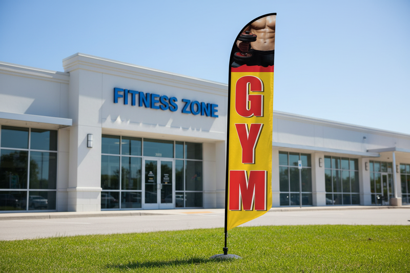 Gym feather flag kit displayed outdoors in grass in front of a fitness center, 11.5ft flag on aluminum pole with ground spike