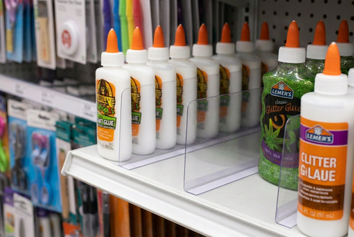 Thin clear PVC shelf divider with a single one-sided adhesive foot separating craft glue bottles on a retail shelf.