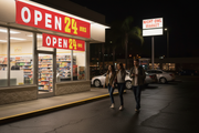 Open 24 Hours vinyl banner hung on the exterior window of a convenience store or gas station at night with glowing storefront lights and customers approaching