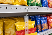 Galvanized pinched S-hook inserted through a round hole in a perforated gondola shelf edge, holding a clear clip strip merchandising strip of snack packets in a retail aisle