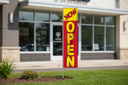 Red-and-yellow Now Open banner flag kit using the original product artwork, planted in landscaped grass outside a storefront with the full pole visible