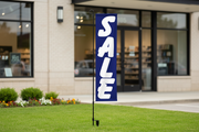 Blue Sale banner flag kit using the original product artwork, planted in landscaped grass outside a storefront with the full pole and stakes visible