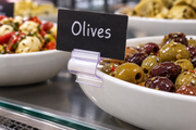 Clear reusable gourmet sign clip gripping the rim of a white deli olive bowl and holding a small black Olives tag in a supermarket case