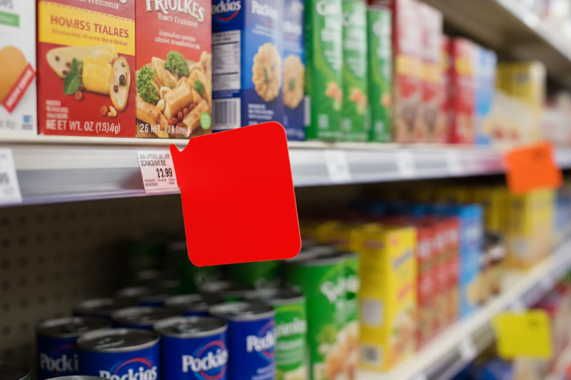 Fluorescent red blank aisle violator flag shelf sign mounted in price channel rail on supermarket shelf, sign in focus with grocery aisle blurred behind - lifestyle in-store display