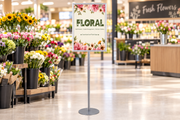 Silver single-sided snap-frame floor stand with a vertical 24 × 36 floral poster in a supermarket floral department.