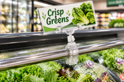 Clear all-plastic spring clip-on sign holder clamped to a chrome cooler rail, swivel knuckle angling a Crisp Greens sign above misted lettuce and bagged salads