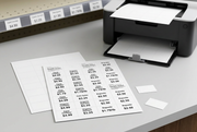 White perforated printable price-tag sheets with sample retail labels and separated tags beside a laser printer, with shelf-channel labels in the background.