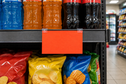 Landscape red-orange fluorescent 8.5 by 5.5 inch shelf price card held by two clear clips on a convenience-store endcap with beverages and snacks