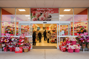 IGA Valentine's Day vinyl banner hung on an IGA supermarket window or entrance with red and pink hearts, romantic seasonal atmosphere, and shoppers browsing floral and candy displays