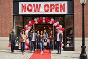 Now Open vinyl banner 8 feet wide hung on the exterior of a retail storefront grand opening with excited customers approaching the entrance and a celebratory atmosphere