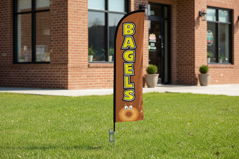 Brown Bagels feather flag using the uploaded artwork, planted in green grass with a visible pointed metal ground spike