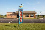 Blue Hot Dogs feather flag planted in a grass strip outside a convenience store in a wide landscape setting, showing the full pole and ground spike