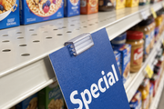 Close-up of a clear one-inch Super Grip shelf sign clip gripping a blue Special paper sign on a grocery shelf