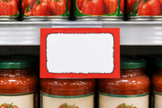 Red starburst 5.5 by 3.5 inch coated cardstock price card in a clear holder on a supermarket shelf with canned goods and jars