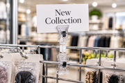 Clear double-clip swivel sign holder clamped to a chrome wire grid fixture rod, upper clip holding a horizontal New Collection card above hanging packaged merchandise