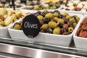 Black oval write-on wipe-off case tag labeled Olives mounted in a clear holder at the front of a supermarket deli olive display
