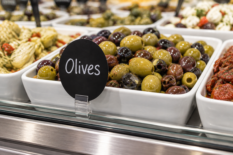Black oval write-on wipe-off case tag labeled Olives mounted in a clear holder at the front of a supermarket deli olive display
