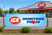 IGA Hometown Proud vinyl banner with red IGA logo and navy lettering mounted by four corner grommets on a fence outside an IGA supermarket