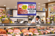 Easter ham hanging sign, approximately 28 x 22 inches, suspended by two top-corner mounts above a staffed supermarket ham counter with Easter display decorations