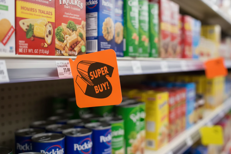 Fluorescent red-orange Super Buy aisle violator flag shelf sign mounted in price channel rail on supermarket shelf, sign in focus with grocery aisle blurred behind - lifestyle in-store display