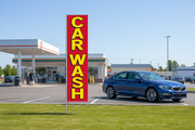 Red and yellow Car Wash banner flag installed in green grass beside a fuel-station car wash with a vehicle, showing roadside advertising use