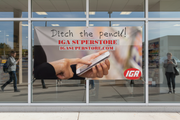 IGA Online Shopping semi-custom vinyl banner 5 feet wide with store name and website URL, hung on an IGA supermarket window or entrance with customers using smartphones