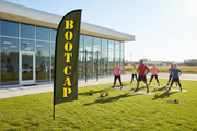 Boot Camp feather flag installed in green grass outside a fitness studio with a workout group, showing outdoor gym promotion use