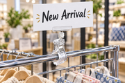 Clear clip-on sign holder with white swivel knuckle clamped to a chrome wire rack rail, top jaw gripping a horizontal New Arrival card in a retail store