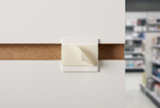 Head-on close-up of a white slatwall sign holder adapter clip with peel-and-stick adhesive tab seated in a slatwall groove, SKU SWSDA