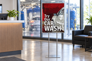 Try Our Car Wash Suds 22 × 28-inch poster with soap-covered car and red brush artwork displayed in a floor stand at a car wash reception.