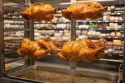 Golden roasted rotisserie chicken with elastic tucker loops holding the legs together, displayed in a grocery store deli rotisserie case.