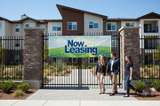 Now Leasing vinyl banner hung on the exterior fence or entrance of a modern apartment complex with prospective renters walking by in bright natural daylight